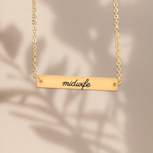 May include: A gold-toned horizontal bar necklace featuring a delicate chain and a rectangular pendant. The center of the bar is engraved with the word midwife in a black, cursive script font, creating a clean and simple jewelry piece.