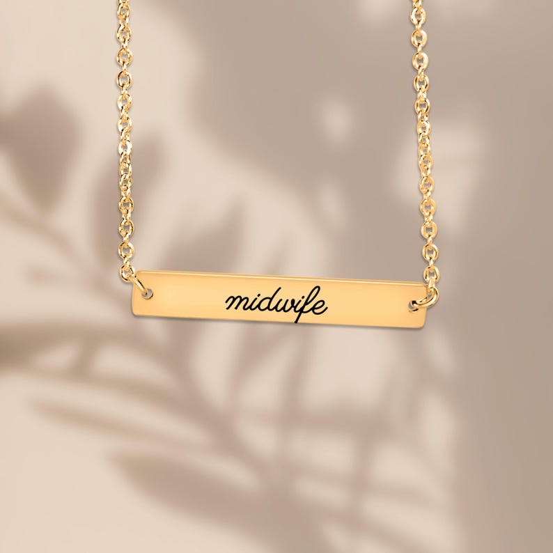 May include: A gold-toned horizontal bar necklace featuring a delicate chain and a rectangular pendant. The center of the bar is engraved with the word midwife in a black, cursive script font, creating a clean and simple jewelry piece.