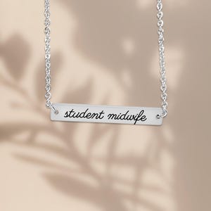May include: A silver-toned horizontal bar necklace featuring the text student midwife engraved in a black cursive script. The pendant is attached to a delicate silver-toned chain with circular links on both the left and right sides.