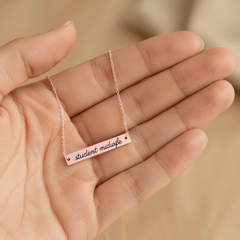 May include: A rose gold-toned bar necklace featuring a delicate chain and a horizontal rectangular pendant. The pendant is engraved with the black cursive text student midwife, centered on the polished metal surface of the bar.