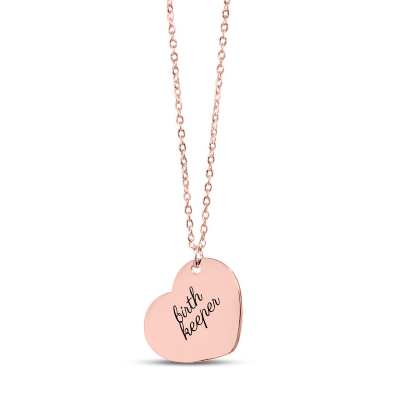 Pu&ograve; includere: Una collana con ciondolo a forma di cuore color oro rosa e una catenina sottile. La superficie liscia e lucida del cuore &egrave; incisa con il testo in corsivo nero birth keeper, centrato sulla parte anteriore del pezzo di metallo.