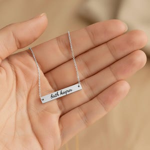 Puede incluir: Un collar de barra horizontal plateado con una cadena delicada y un colgante rectangular grabado con el texto birth keeper en una fuente cursiva negra. La superficie met&aacute;lica tiene un acabado liso y pulido en toda la pieza.