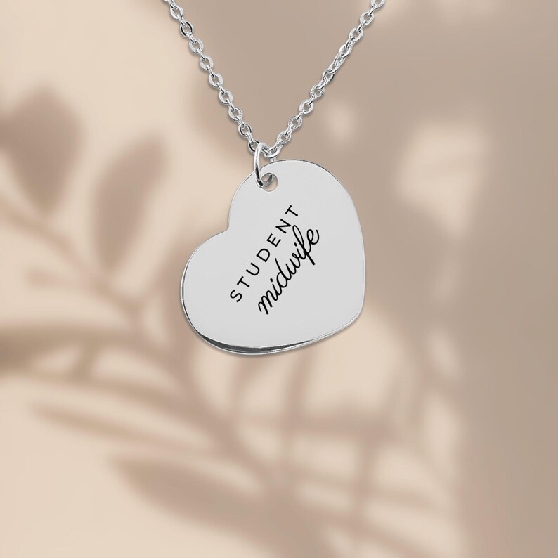 May include: A silver-toned heart-shaped pendant necklace featuring the engraved text "STUDENT midwife" in a combination of block and script lettering, suspended from a delicate silver-toned chain link necklace.