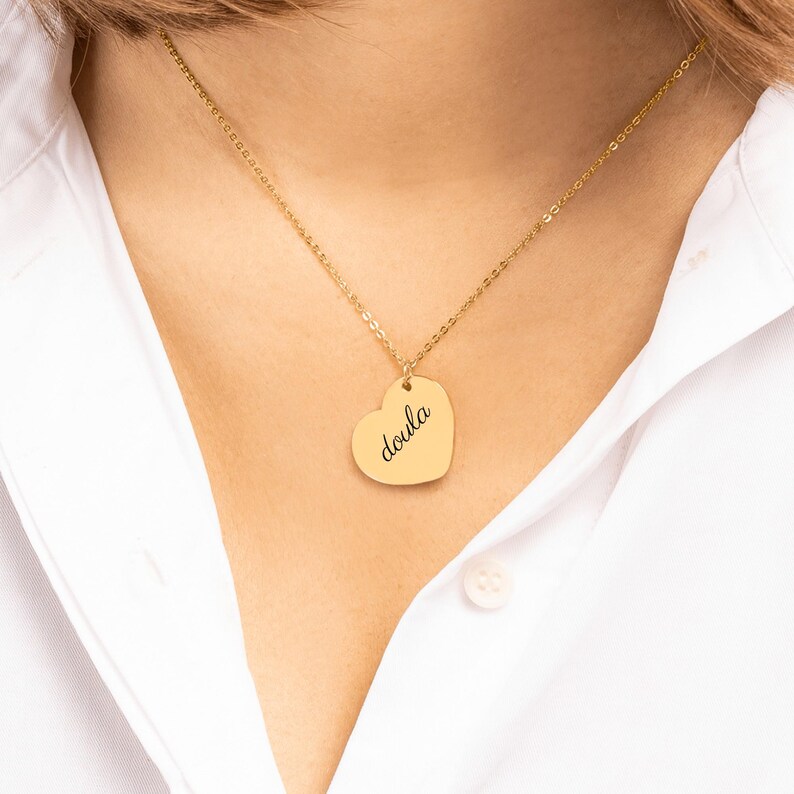 Puede incluir: Un collar con colgante en forma de coraz&oacute;n dorado que presenta la palabra doula grabada en letra cursiva negra. El colgante cuelga de una delicada cadena dorada, descansando sobre una camisa blanca de cuello con un bot&oacute;n visible.