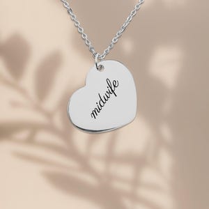 May include: A silver-colored heart-shaped pendant necklace featuring the word midwife engraved in a black cursive script. The pendant hangs from a delicate silver-colored chain link necklace against a neutral background.