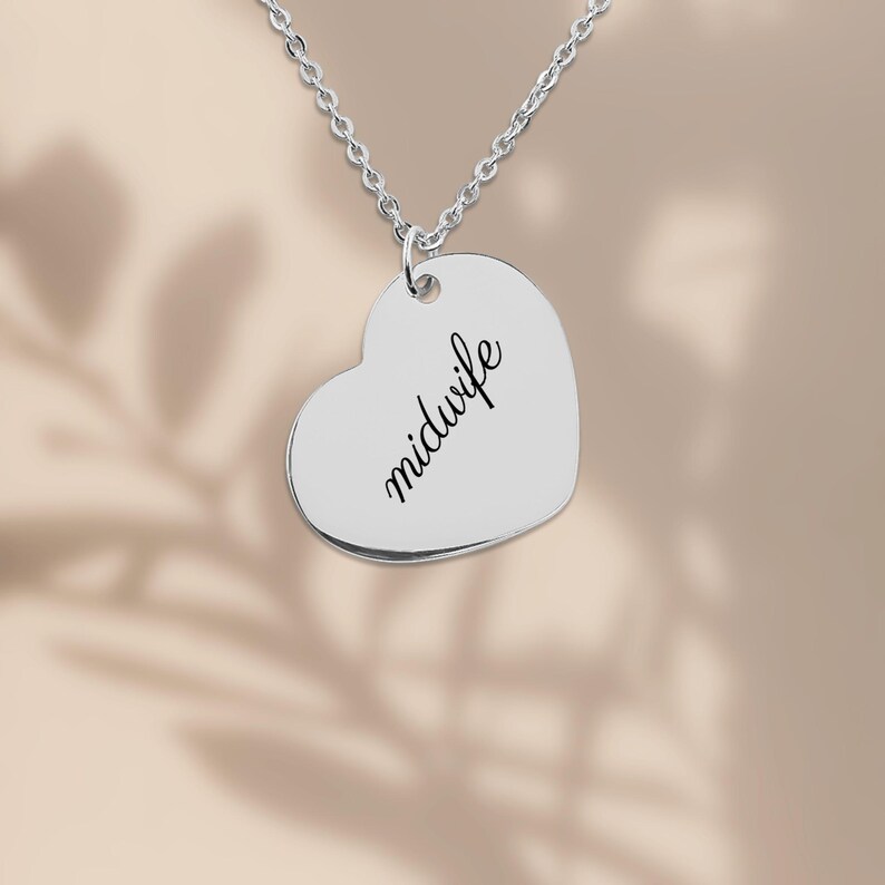 May include: A silver-colored heart-shaped pendant necklace featuring the word midwife engraved in a black cursive script. The pendant hangs from a delicate silver-colored chain link necklace against a neutral background.