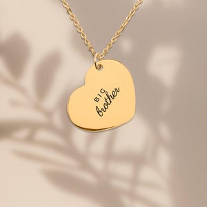 Op de afbeelding: Een goudkleurige hartvormige hanger aan een ketting met de gegraveerde tekst "BIG brother" in een combinatie van blokletters en cursief schrift. De hanger hangt aan een fijne goudkleurige ketting met een gepolijste, reflecterende afwerking.