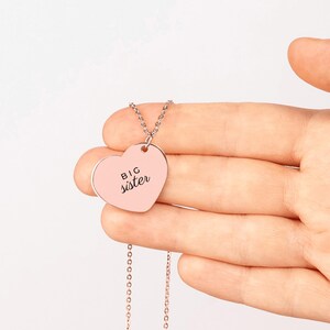 May include: A rose gold heart-shaped pendant necklace featuring the text BIG sister engraved in black. The pendant is attached to a delicate matching rose gold chain, creating a simple and elegant piece of jewelry for everyday wear.
