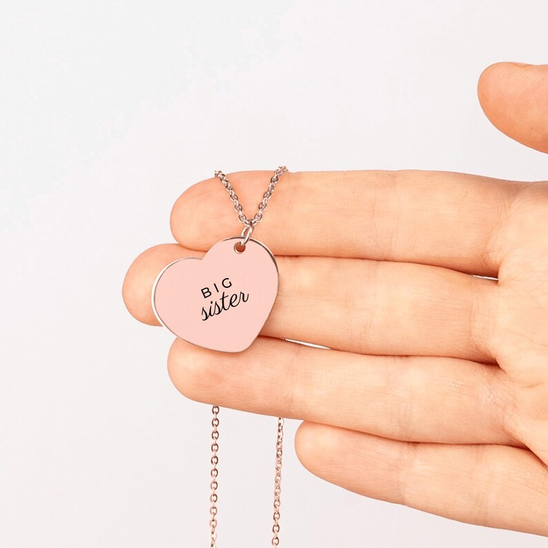 May include: A rose gold heart-shaped pendant necklace featuring the text BIG sister engraved in black. The pendant is attached to a delicate matching rose gold chain, creating a simple and elegant piece of jewelry for everyday wear.