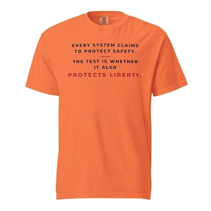 Pode incluir: Uma t-shirt laranja de manga curta com texto preto que diz: EVERY SYSTEM CLAIMS TO PROTECT SAFETY. THE TEST IS WHETHER IT ALSO, seguido pela palavra PROTECTS LIBERTY. em vermelho. A t-shirt tem gola redonda e um corte padrão.