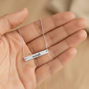 May include: A silver-toned horizontal bar necklace featuring the word midwife engraved in a black cursive script. The pendant is attached to a delicate silver-toned chain that extends from both sides of the rectangular metal bar.
