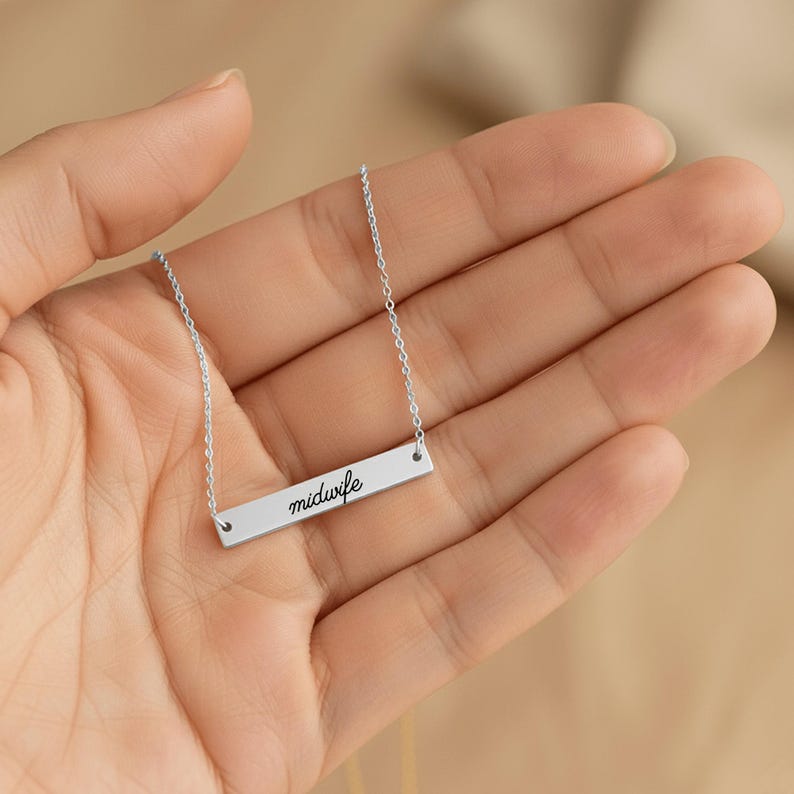 May include: A silver-toned horizontal bar necklace featuring the word midwife engraved in a black cursive script. The pendant is attached to a delicate silver-toned chain that extends from both sides of the rectangular metal bar.