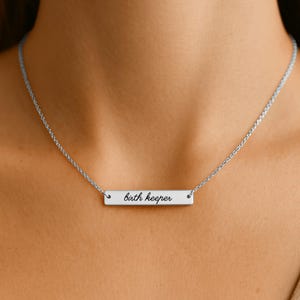 Puede incluir: Un collar con una barra horizontal plateada que presenta el texto grabado "birth keeper" en una fuente cursiva. El colgante est&aacute; suspendido de una delicada cadena plateada que descansa alrededor del cuello, con un dise&ntilde;o limpio y sencillo.