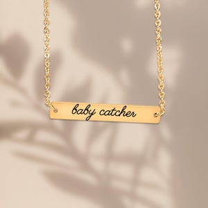 May include: A gold-toned bar necklace featuring a delicate chain and a horizontal rectangular pendant. The pendant is engraved with the text "baby catcher" in a black, cursive script font, centered on the polished metal surface.
