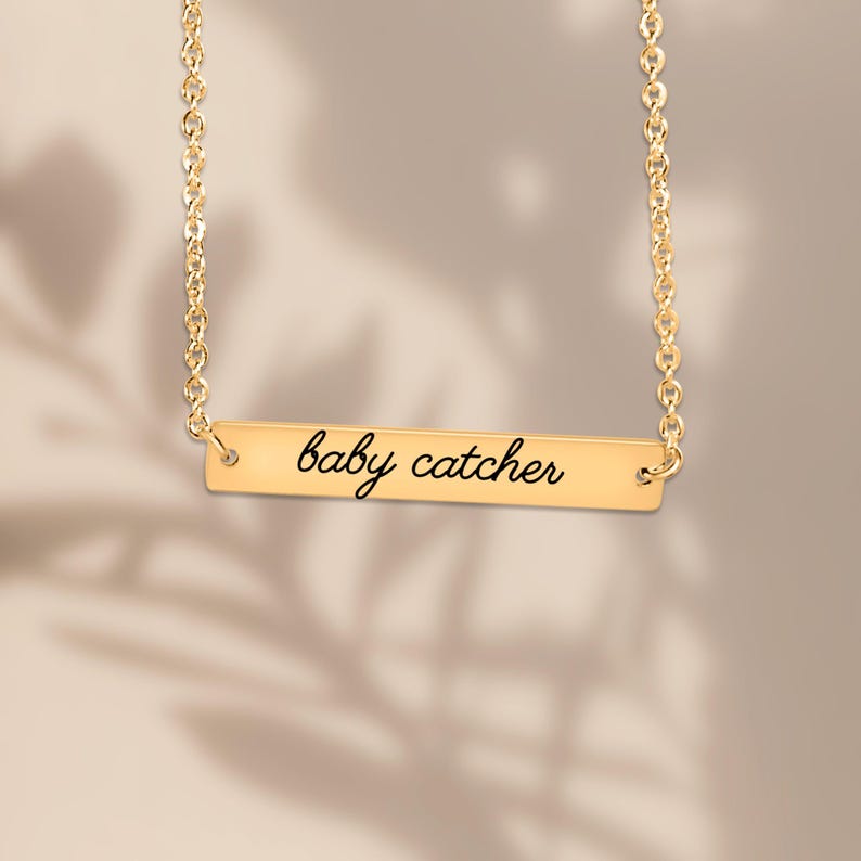 May include: A gold-toned bar necklace featuring a delicate chain and a horizontal rectangular pendant. The pendant is engraved with the text "baby catcher" in a black, cursive script font, centered on the polished metal surface.