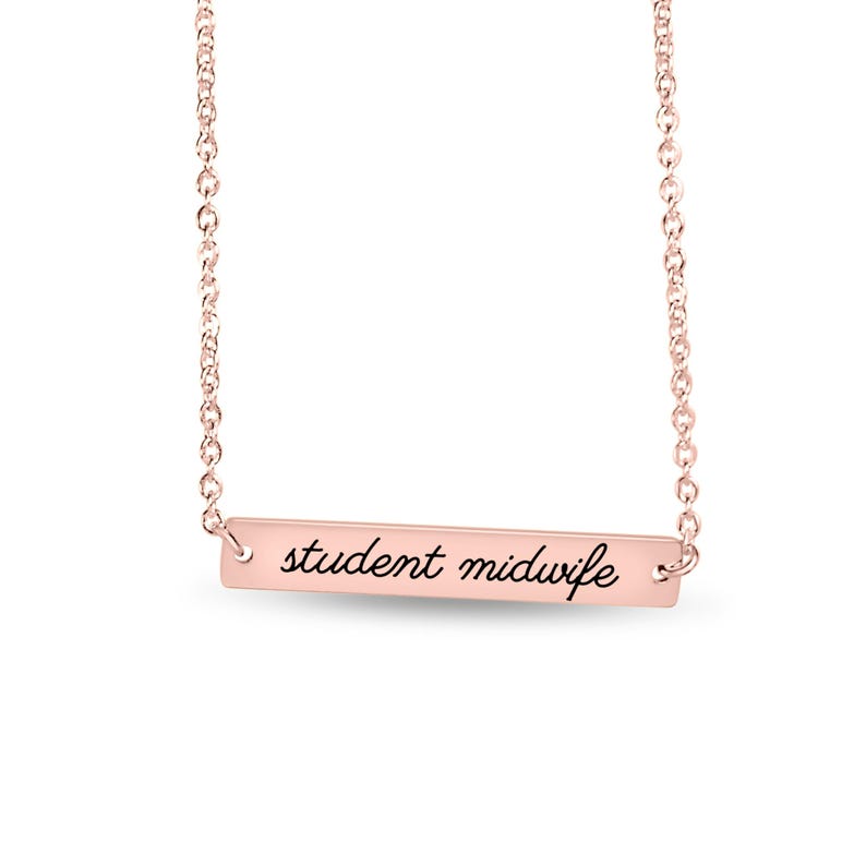 May include: A rose gold-toned bar necklace featuring a delicate chain and a horizontal rectangular pendant. The pendant is engraved with the black cursive text "student midwife" centered across its smooth, polished metallic surface.