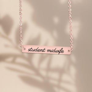 May include: A rose gold-toned bar necklace featuring a delicate chain and a horizontal rectangular pendant. The pendant is engraved with the black cursive text "student midwife" centered on its smooth, polished metallic surface.
