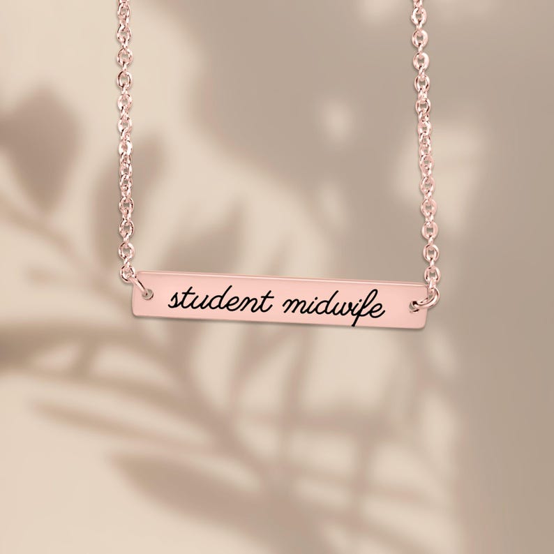 May include: A rose gold-toned bar necklace featuring a delicate chain and a horizontal rectangular pendant. The pendant is engraved with the black cursive text "student midwife" centered on its smooth, polished metallic surface.