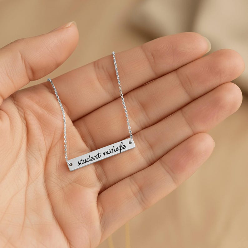 May include: A silver-toned bar necklace featuring a delicate chain and a rectangular pendant engraved with the black cursive text student midwife. The metal bar is polished and reflects light, suspended from a thin, matching silver-colored link chain.
