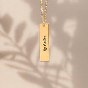 Puede incluir: Un collar con colgante rectangular vertical en tono dorado y cadena fina. La superficie met&aacute;lica lisa y pulida del colgante est&aacute; grabada con el texto en cursiva negra big brother, centrado verticalmente en la cara frontal.