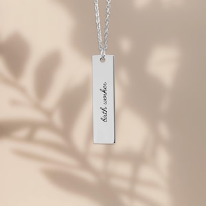 May include: A silver-toned vertical bar pendant necklace featuring the engraved text "birth worker" in a black script font. The rectangular metal charm hangs from a matching silver-toned chain, creating a clean and simple jewelry piece.