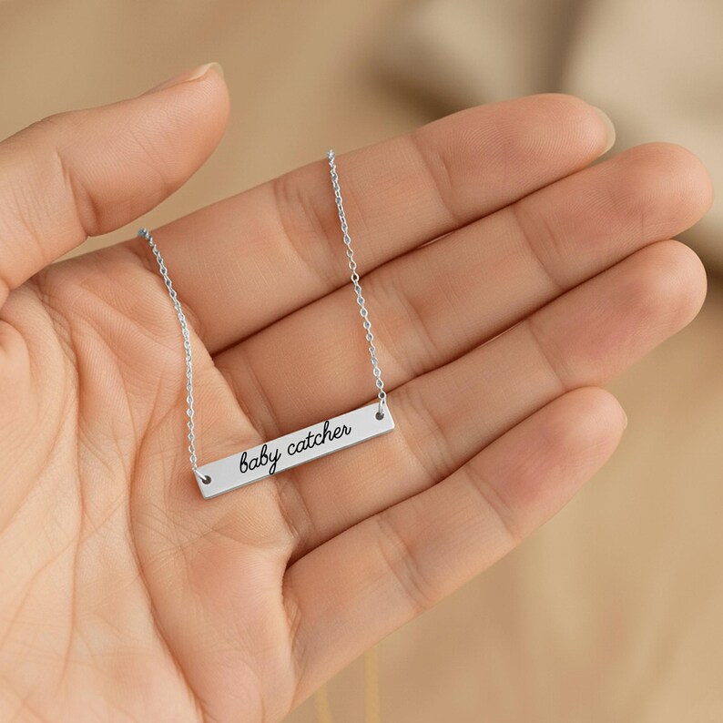 May include: A silver-toned horizontal bar necklace featuring the text baby catcher engraved in a black cursive script. The pendant is suspended from a delicate silver-toned chain, showcasing a clean and simple design for everyday wear.
