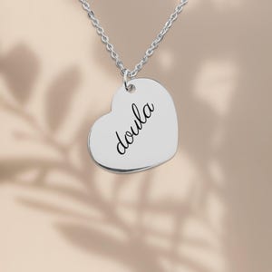 Puede incluir: Un collar con colgante en forma de coraz&oacute;n de color plateado que presenta la palabra doula grabada en una tipograf&iacute;a cursiva negra. El colgante cuelga de una delicada cadena plateada, creando una pieza de joyer&iacute;a sencilla.