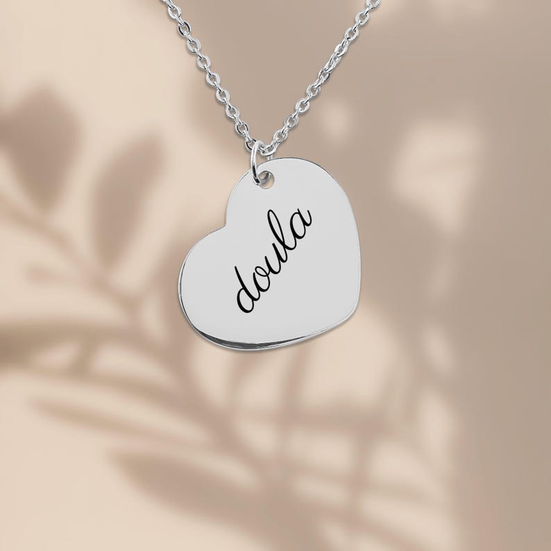 Puede incluir: Un collar con colgante en forma de coraz&oacute;n de color plateado que presenta la palabra doula grabada en una tipograf&iacute;a cursiva negra. El colgante cuelga de una delicada cadena plateada, creando una pieza de joyer&iacute;a sencilla.