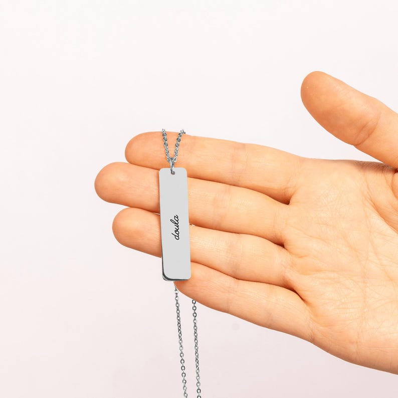 May include: A silver-toned metal bar pendant necklace featuring a delicate chain and a rectangular charm. The charm is engraved with the word doula in a black cursive script font, centered vertically on the smooth, polished surface.