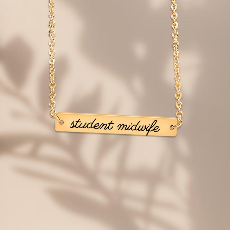 May include: A gold-toned necklace featuring a horizontal rectangular bar pendant. The bar is engraved with the black cursive text student midwife. The pendant is suspended from a delicate gold-toned chain with circular links.