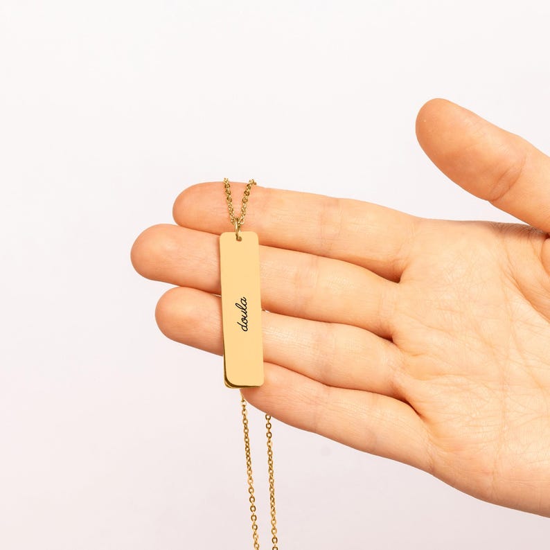May include: A gold-toned vertical bar pendant necklace featuring the word doula engraved in a black script font. The rectangular metal charm is suspended from a delicate matching gold-toned chain, creating a simple and clean jewelry piece.