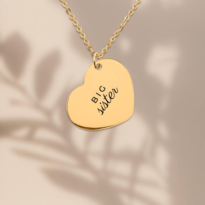 May include: A gold-toned heart-shaped pendant necklace featuring the engraved text BIG sister in a combination of block and script lettering. The pendant hangs from a delicate gold-toned chain, showcasing a smooth, polished metallic finish.