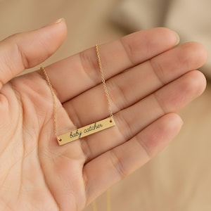 May include: A gold-toned bar necklace featuring a delicate chain and a horizontal rectangular pendant. The pendant is engraved with the text baby catcher in a black cursive script font, centered on the polished metal surface.