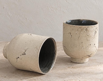 Può includere: Due tazze in ceramica con un esterno testurizzato color bianco sporco con macchie scure irregolari e un interno liscio grigio scuro. Una tazza è posizionata in verticale su una piccola base circolare, l'altra è appoggiata su un lato.