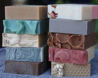 May include: Two stacks of rectangular bar soap in various colors and textures. The left stack features solid tan, teal, speckled cream, marbled gray, and dark brown bars. The right stack includes white with dried petals, solid gray, brown, pink, and honey-patterned bars.