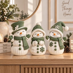 May include: Three white snowman figurines with green accessories. The left snowman wears a top hat and scarf. The center snowman has a knit hat and scarf. The right snowman wears a knit cap with a pom-pom and scarf. All have orange carrot noses.