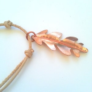May include: A copper-coloured leaf pendant with a natural, organic design. The pendant is attached to a tan, knotted cord necklace. The leaf has detailed veins and a slightly textured surface, creating a rustic and earthy aesthetic. The necklace is suitable for everyday wear.