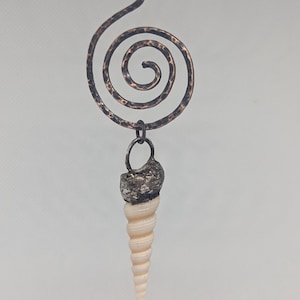 May include: A pendant featuring a hammered copper spiral attached to a dark metal ring, which holds a long, cream-coloured, conical seashell. The copper spiral has a textured, dark finish, and the shell is attached to the ring with a dark metal cap.