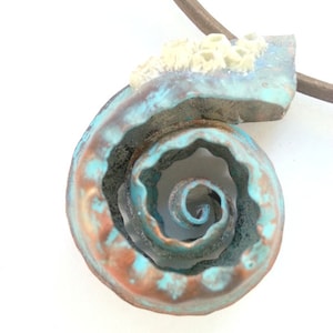 May include: A turquoise and copper coloured spiral shell pendant necklace. The shell is made of metal and has a textured surface. The necklace is made of brown leather cord.