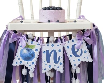 May include: A white high chair decorated for a first birthday. The chair has a banner that says "one" with blueberry illustrations. A small cake with blueberries sits on the tray. The chair is decorated with purple and blue ribbons.