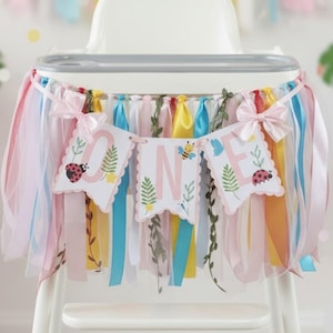 May include: A high chair decorated for a first birthday. The chair has a banner reading "ONE" and colourful ribbons. The banner features ladybirds, bees, and floral accents. Pastel-coloured ribbons and tulle create a festive look, perfect for a party.