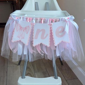 May include: A white high chair decorated with a pink and white tulle skirt and banner that reads "one". The banner has pink bows and scalloped edges. The high chair has a metal frame and is set on a light wood floor.