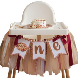 Puede incluir: Una trona blanca decorada con una falda de tul beige y un cartel con la palabra one y una ilustración de una tarta. El cartel está adornado con cintas de satén rojo oscuro. Un pequeño pastel blanco descansa sobre la bandeja.