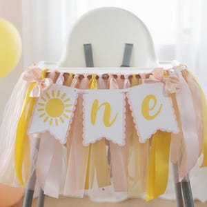 May include: A high chair banner featuring three white scalloped pennants with yellow text and a sun graphic spelling out the word one. The banner is decorated with long strips of yellow, peach, and white tulle and satin ribbons on each side.