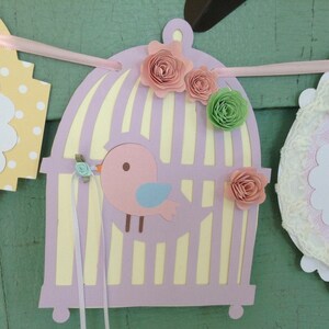 May include: A pink and purple birdcage banner with a pink bird inside. The banner has yellow polka dots, white lace, and pink and green paper flowers.