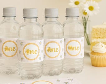 Puede incluir: Cuatro botellas de agua de plástico transparente con tapas grises, cada una con una etiqueta con un sol amarillo y la palabra "one". Las botellas son parte de un tema de fiesta, con una pancarta, globos y pastel a juego.