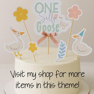 以下が含まれることがあります： 「One Silly Goose」をテーマにしたデコレーションケーキ。パステルカラーのガチョウと花のトッパー、ピンクのリボンで飾られています。ケーキには「Visit my shop for more items in this theme!」と書かれています。