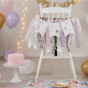 May include: A white high chair decorated with a banner that spells "one" with illustrations of fairies. The chair is adorned with pink, white, and lavender ribbons. A pink cake sits on a white cake stand, surrounded by balloons in gold, pink, and lavender.