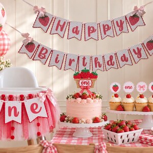 May include: A strawberry-themed first birthday party set featuring a Happy Birthday banner, a high chair tutu with a one banner, a cake topper with the word one, and cupcake toppers with strawberries, bows, and the word one on a table.