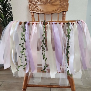 May include: A wooden chair decorated with a tulle skirt and ribbons in shades of pink, white, and lavender. Green vine accents are interspersed among the ribbons, creating a whimsical and festive look. The chair has a decorative back.